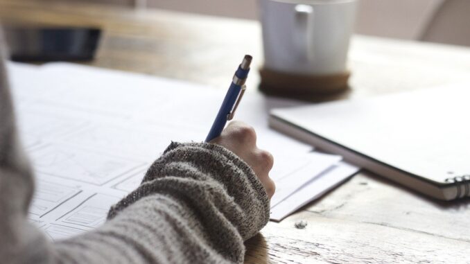 Ruka drží pero a píše na stole, kde je položena káva nabílý papír.