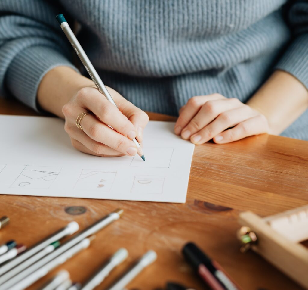 žena sedící za stolem, kreslí - píše na čtvrtku bílého papíru. kolem má rozsypané psací potřeby