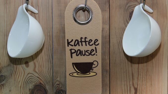 Tři háčky na stěně, dva s pověšenými bílými hrnečky, jeden s cedulkou na které je napsané kaffee pause.