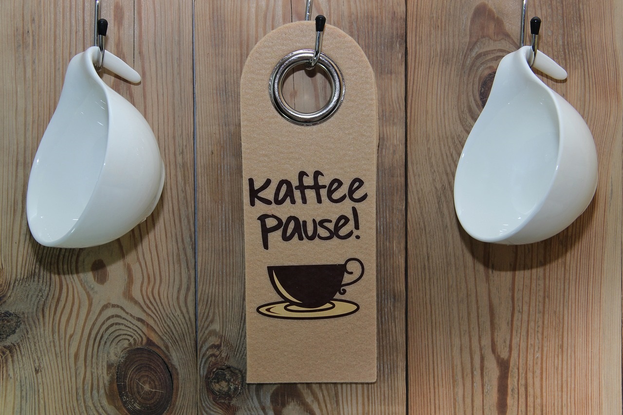 Tři háčky na stěně, dva s pověšenými bílými hrnečky, jeden s cedulkou na které je napsané kaffee pause.
