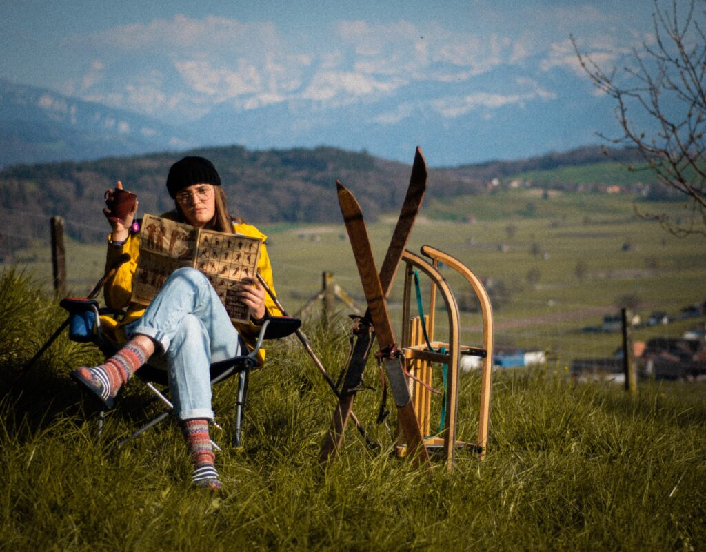 Žena sedí v křesílku na trávě, vedle sebe lyže a sáňky, a čte si.