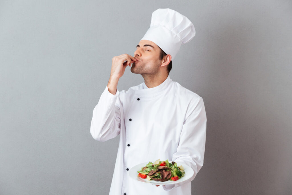 Muž v kuchařské čepici a s talířem v ruce gestem naznačuje, že jídlo je vynikající.
