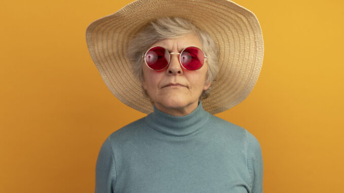 confused old woman wearing blue turtleneck sweater beach hat and sunglasses looking at camera isolated on orange background