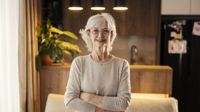 Portrait of confident senior woman with arms crossed smiling at camera at cozy home.