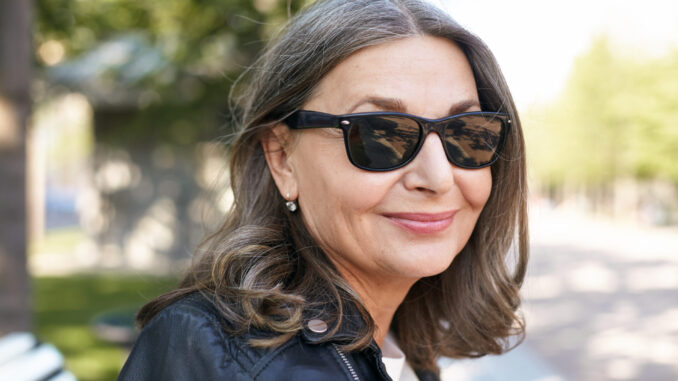 Close up portrait of modern stylish mature fifty year old grandmother with neat hairstyle, posing outdoors wearing black leather jacket and sunglasses, looking at camera with joyful happy smile