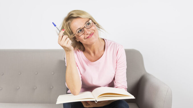 mature-woman-sitting-sofa-holding-pen-open-book-daydreaming