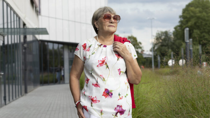 Real Modern Senior Woman In Sunglasses Walks Near Shopping Mall. Green Trees, Park on Background. Portrait Of Happy Elderly Female, Enjoying Retirement Life In City. Authentic Lifestyle Horizontal
