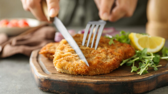 Woman eating delicious schnitzel with microgreens and lemon at grey table, closeup