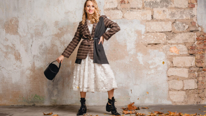 attractive stylish blonde woman in checkered jacket coat walking against wall in street, autumn fashion trend, wearing white lace dress, boots, handbag, suitcases, leaves on the ground, urban style