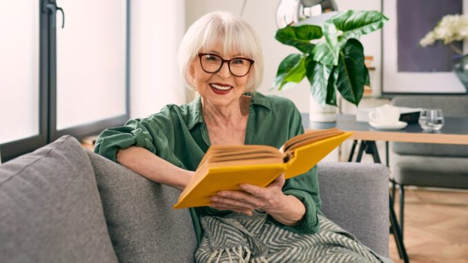 cheerful-senior-woman-sitting-couch-reading-book-home