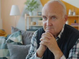 Elderly man sitting on a couch with hands clasped.