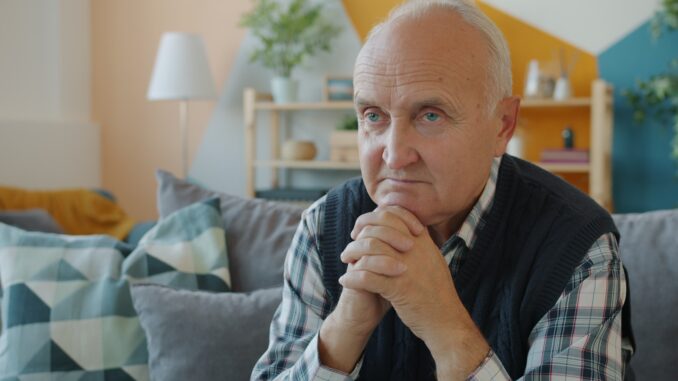 Photo by Vitaly Gariev Elderly man sitting on a couch with hands clasped.